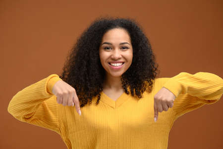 Charming young african-american woman points index fingers down, promoting novelty isolated on brown...