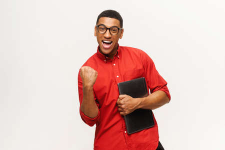 Excited overjoyed African-American young man in red casual shirt celebrating good news or winning, expresses happiness, latin guy with laptop screaming yes in sign of triumphの写真素材