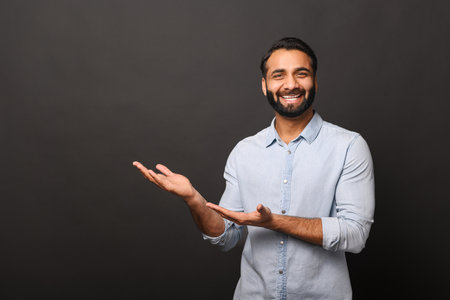 Happy Indian bearded man in casual shirt points hands aside at empty space, hispanic man advertising novelty standing isolated on black, showing with palms sidewayの写真素材