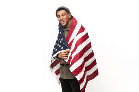 Smiling and cheerful guy protesting against racism, isolated on white wall. Happy latin man fighter for rights wrapped at United States flagの写真素材