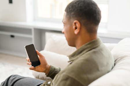 Side view of the multiracial man sitting on sofa at home and holding smartphone. Hispanic guy reading news online and spending leisure time in social networksの写真素材