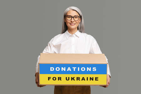 Smiling gray-haired woman encourages to share things with Ukrainian refugees, middle-aged Asian lady...