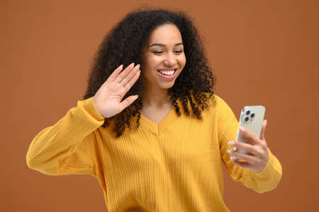 Joyful African American young woman using smartphone for video call stands isolated on brown, video chatting, speaking on mobile phone via video and smiling. Video call conceptの写真素材