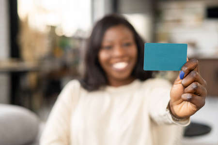 Selective focus at card in hand of happy smiling African-American woman, cheerful lady showing empty gift card, presenting banking credit card, copy spaceの写真素材