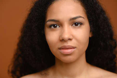 Young african woman looking at the camera, posing over brown background. Well-looking lady with naked shoulders, studio shot. Natural beauty and skincare conceptの写真素材