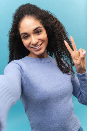 Happy carefree African-American young woman with curly hair taking selfie on the smartphone, gesturing, making content for social medias, isolated on blueの写真素材