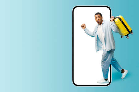 Excited African-American guy carrying yellow suitcase running inside huge mobile phone, scared multiracial man fears does not take advantage of a good offer, hurrying. App for travelersの写真素材
