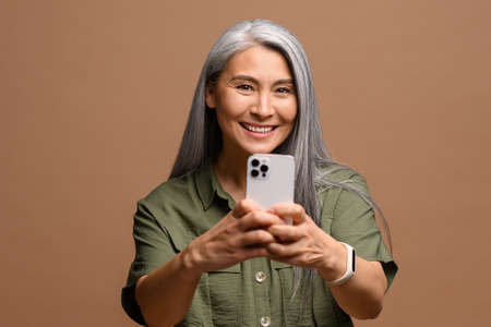 Cheerful attractive senior woman holding smartphone, enjoying chatting in social networks, shooting...