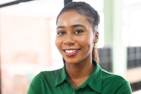 Close-up portrait of confident african-american female employee standing in modern office space, cheerful attractive black businesswoman in green shirt looks at the camera with light friendly smileの写真素材