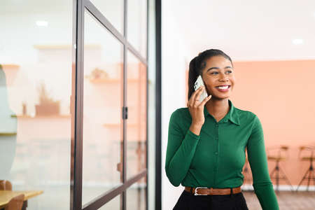 Confident African-American businesswoman talks phone walking down office hall, cheerful friendly female employee speaks on mobile phone with clients, chatting on smartphoneの写真素材