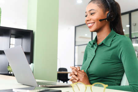 Young cheerful african-american woman using headset for online communication sitting at modern office. Pleasant female operator speaking with customers or coworkers on the distanceの写真素材