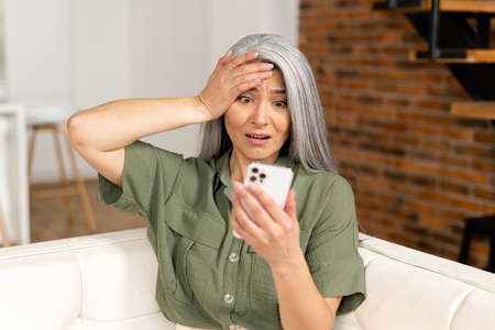 Worried senior woman staring at a smartphone screen at home, upset retire female sitting on the couch looks at mobile phone with vexation, read terrible news or received bad analysis resultsの写真素材