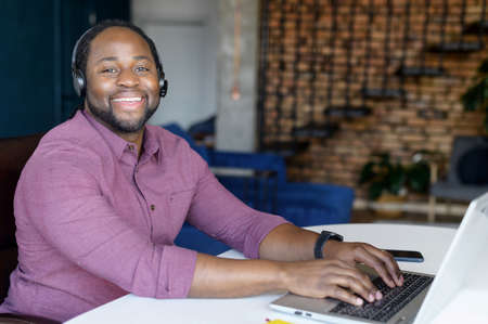 Friendly and positive African-American guy wearing headset using laptop computer working remotely...