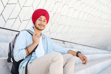 Modern male Indian student with backpack wearing red national turban pagri sitting on the steps outdoors, hindu guy in traditional headwrap looking at the camera with pleasant smileの写真素材