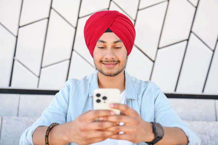 Portrait of cheerful young Indian guy sending message on mobile phone outdoors. Happy hindu man in...