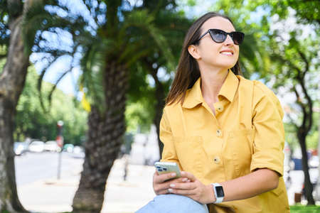 Attractive cheerful woman using smartphone sitting in the park, smiling female student in sunglasses...
