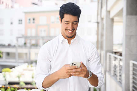 Handsome hispanic guy is using smartphone for messaging, texting, young dark-haired man in smart casual white shirt using smartphone outdoors, typing a message, chatting in the networks, scrollingの写真素材