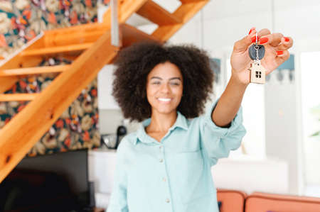 Selective focus at keys with cute keychain little house showing by young happy woman, excited female student moved in own dwelling, african-american relocated, moving into a new house, got a mortgageの写真素材