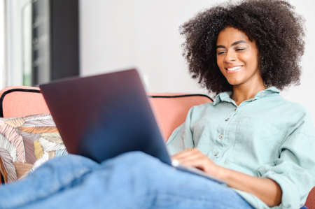 African young woman student or freelancer in casual wear sitting on the couch and using laptop, working on the project, looking at the screen with pleasure smile, websurfing or shopping onlineの写真素材