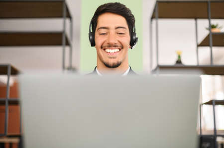 Young latin businessman with headset sitting at the desk and talking by video with new customers or investors, full of ideas, performing online, taking part webinar podcast on laptop, listening courseの写真素材