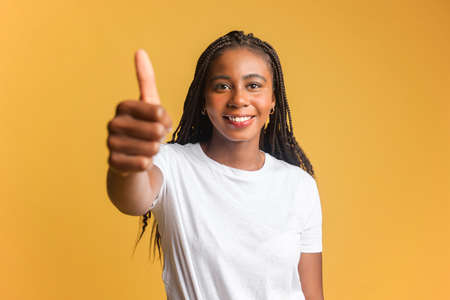 Like, excellent job. Pleased woman with cool hairstyle smiling at camera and gesturing thumbs up, approval hand sign, satisfied with result. Studio shot isolated on yellow backgroundの写真素材