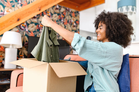 Positive young black woman standing with carton box and taking out new clothes, while feeling curious about ordered item from online store. Smiling client satisfied with fast courier delivery serviceの写真素材