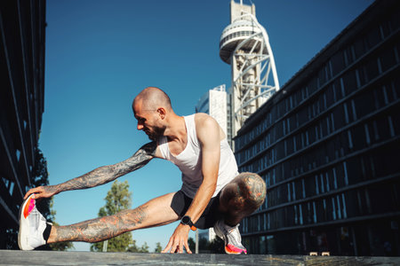 Young muscular tattooed man doing exercises, stretching early in the morning, bald bearded guy in sportswear stretching legs before joggingの写真素材