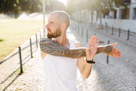 Young muscular tattooed man doing exercises, stretching early in the morning, bald bearded guy in sportswear stretching arms before joggingの写真素材