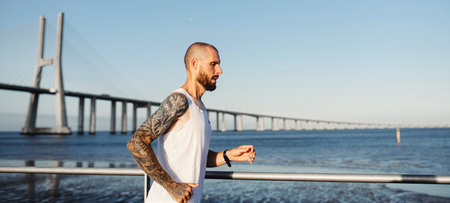 Tattooed young man jogging on the rivercoast on the sunset. Banner with athletic guy doing cardio workout outdoorsの写真素材