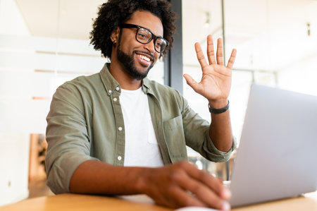 In touch on the distance. Confident and happy african-american male employee using online platform for virtual communication with coworkers, waving hand at webcam, making video call indoorの写真素材