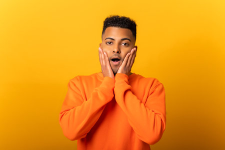 Serious shocked man holding hands on face and screaming looking at camera with open mouth, shocked bemused with news. Indoor studio shot isolated on yellow backgroundの写真素材