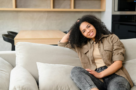 Headshot of beautiful smiling serene young woman in casual shirt resting on the sofa at cozy apartment, charming multiracial girl looking at the camera with wide smileの写真素材