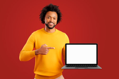 Positive smiling man freelancer with beard showing and holding laptop with empty screen, satisfied with teleworking, likes his job. Indoor studio shot isolated on red backgroundの写真素材