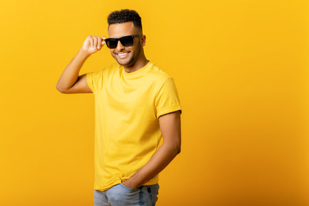 Overjoyed Indian man wearing sunglasses looking at the camera, smiling positively, being in high spirit. Carefree hispanic guy in yellow t-shirt isolated, having summer moodの写真素材