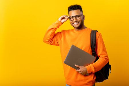 Smart inteligente indian male employee or freelancer man holding laptop and looking at camera with happy smile, isolated on yellow background, arab student with backpack adjusting his glassesの写真素材