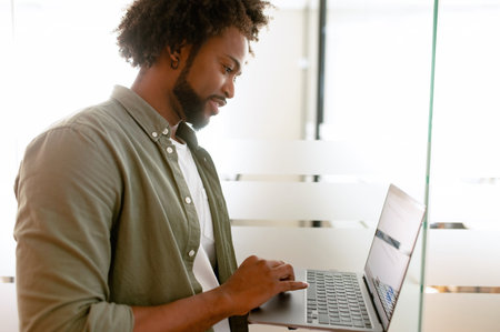 Optimistic ethnic man in casual wear using laptop sitting in modern open space office, young african-american male employee replying emails, working on a new project, side viewの写真素材