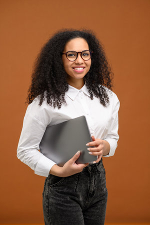 Happy young african woman in formal wear standing isolated over brown background, carrying laptop computer and looking at the cameraの写真素材