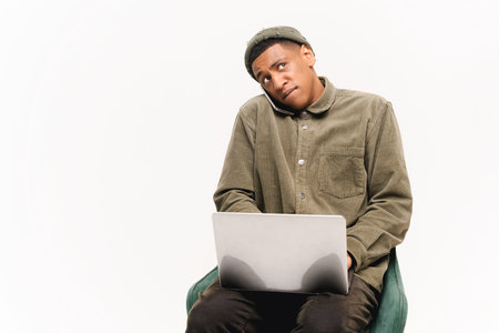 Busy young african-american man sitting with laptop computer and talking by smartphone, concentrated guy having important phone conversation isolated over white backgroundの写真素材