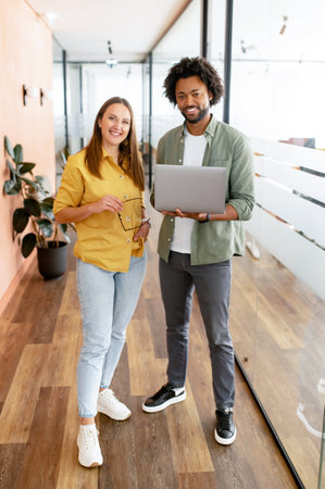 Two coworkers man and woman is using laptop, standing at the office. They holding laptop, looking at camera and smiling, full lengths, friendly office atmosphere and teamwork conceptの写真素材