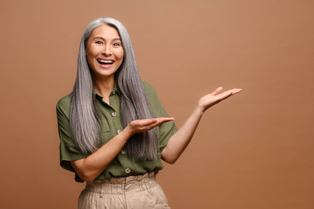 Excited grey haired 50s asian woman pointing away with both hands on blank copy space, showing place for your advertisement isolated over brown backgroundの写真素材