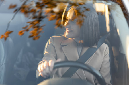 View through the windshield at cheerful young woman sitting in the car and holding steer wheel,...