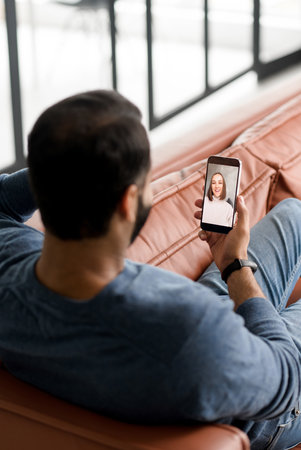 Vertical shot of Indian man resting using mobile dating app, holding phone and talking with woman via video connection, back view dark-haired hispanic latin guy making video call to femaleの写真素材