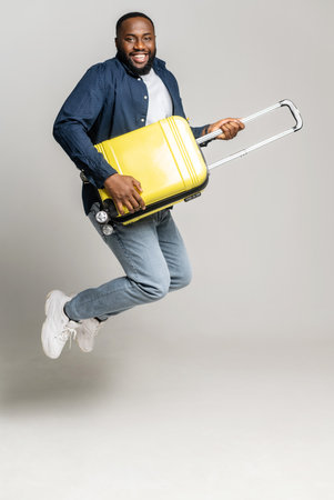 Joyful African-American guy jumping with a yellow suitcase, plays on a bag like a guitarist, fooling around, a black man with luggage looking forward a trip, isolated on gray, full length, verticalの写真素材