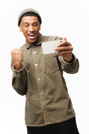 Excited young man playing video game on the smartphone isolated on white background, hispanic gambling guy spends leisure time with video games, celebrating victory, creaming yesの写真素材