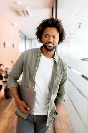 Portrait of an stylish african-american businessman standing in the modern office and posing against the glass partition with the laptop in hand, curly male employee looking at the camera, verticalの写真素材