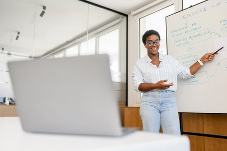 Confident young african woman conducts webinar, business training online. Online coach stands near...