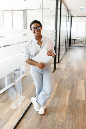 High-motivated female employee carrying laptop in modern office hallway, showcasing positive and...