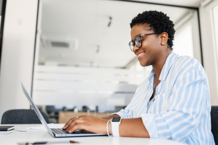 Optimistic african-american female office employee using laptop in modern coworking space, businesswoman in casual wear working on computer, watching at the screen and smiling, side viewの写真素材