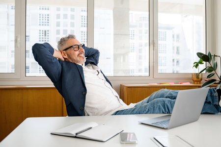Satisfied with well done project mature gray-haired 60s businessman sits in a confident pose with his feet on the table and relaxes, leaned back in the chair, holding his hands behind his headの写真素材