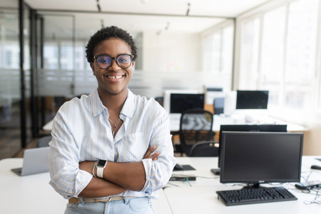 African-American female office employee with short hair standing with arms crossed in contemporary co-working office space, multiracial woman looking at camera. Diverse work team conceptの写真素材